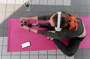 Woman with prosthetic leg stretching on a yoga mat outdoors while wearing headphones, with smartphone and water bottle nearby — inclusive fitness and adaptive yoga