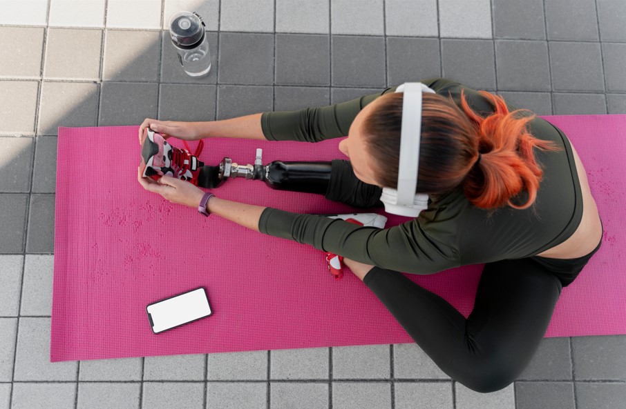 Woman with prosthetic leg stretching on a yoga mat outdoors while wearing headphones, with smartphone and water bottle nearby — inclusive fitness and adaptive yoga